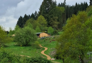 Scenic view of grassy area, trees, winding path and wooden cabin at Campsite Junora, France under cloudy skies.