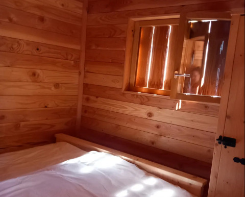 Vista interna di uno chalet in legno con finestra aperta e letto, presso Self-sufficient, luxury eco-chalet, Campeggio Junora, Francia.