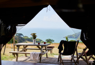 View from inside a tent towards a picnic table, ocean scenery, and hills at BoerenBed Fountain Violet Farm.