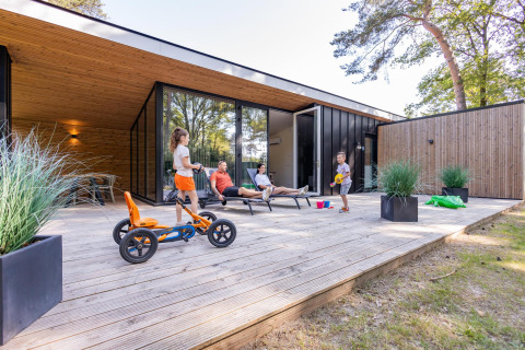 Gezin ontspant op het houten terras van Forest Cottage, spelende kinderen, relaxende volwassenen buiten.