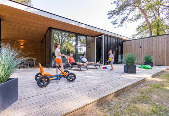 Famiglia si rilassa sulla terrazza in legno del Forest Cottage, bambini giocano e adulti si riposano.