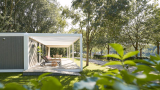 Udendørs terrasse ved Forest Cube-lodge med liggestole og spisebord blandt grønne træer i solskin.