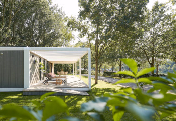 Udendørs terrasse ved Forest Cube-lodge med liggestole og spisebord blandt grønne træer i solskin.
