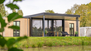 Moderne træhytte ved søen, Forest Lodge, omgivet af grønne træer og komfortable terrassemøbler udenfor.