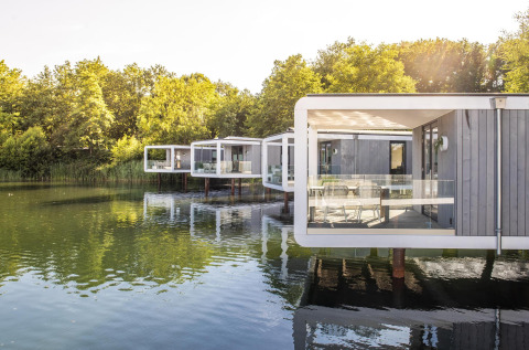 Moderne Water Cube-lodges op palen boven een rustig meer, omgeven door bomen en waterreflecties.