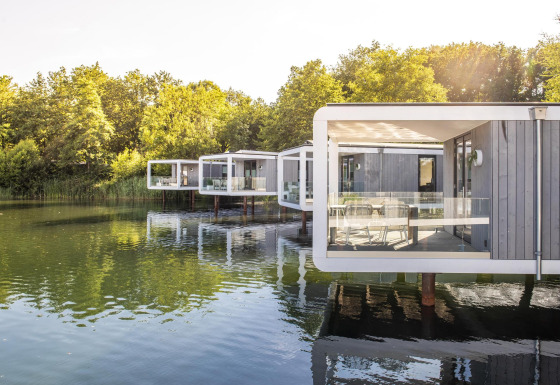Moderne Water Cube-lodges op palen boven een rustig meer, omgeven door bomen en waterreflecties.