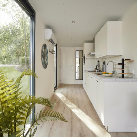 Moderne lodgekeuken met witte kasten, groot raam, houten vloer en uitzicht op een groene tuin.