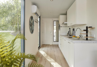 Moderne lodgekeuken met witte kasten, groot raam, houten vloer en uitzicht op een groene tuin.