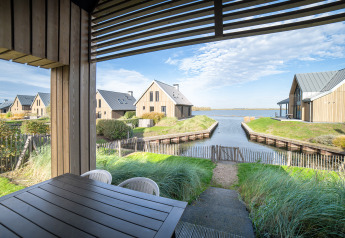Blick von einer überdachten Terrasse auf Wasser, Gras und Holzvillen im Oesterdam Resort in den Niederlanden.