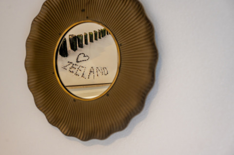 Runder Spiegel mit goldenem Rahmen, reflektiert Flaschenkunst und das Wort 'ZEELAND' in Villa Oesterdam.