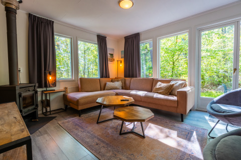 Sala de chalet finlandés en De Wiltzangh, Países Bajos, con sofá esquinero, ventanales y vistas al bosque.