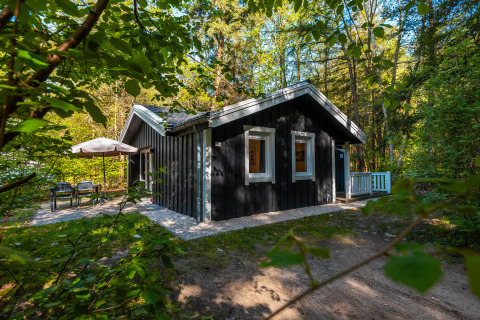 Sort træhytte ved navn Stuga Hot tub omgivet af skov, med udendørs siddeområde og parasol.
