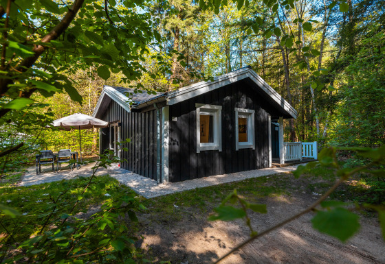 Sort træhytte ved navn Stuga Hot tub omgivet af skov, med udendørs siddeområde og parasol.