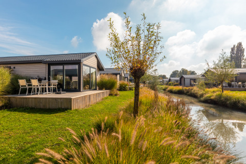 Tiny house Velthorst avec terrasse, au bord d’un canal, pelouse, arbres et ciel ensoleillé.