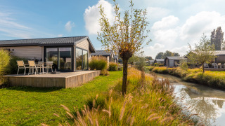 Velthorst tiny house met houten terras aan een gracht, grasveld, bomen en een zonnige blauwe lucht.