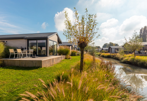 Tiny house Velthorst avec terrasse, au bord d’un canal, pelouse, arbres et ciel ensoleillé.