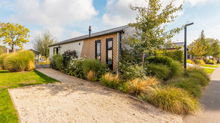 Moderno lodge Berkel circondato da verde e piante ornamentali, con vialetto di ghiaia al sole.