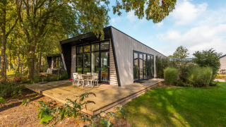 Lodge moderne avec grandes baies vitrées et terrasse en bois entouré de verdure, coin repas extérieur.