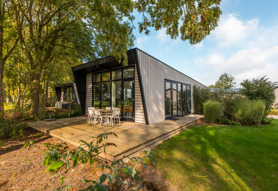 Modern lodge with large windows and a wooden deck, surrounded by greenery and outdoor dining area.