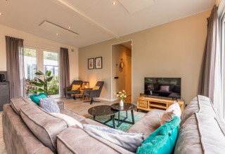 Modern living room in 'Just Nature' lodge at Holiday park Aan de Maas, Netherlands, with sofa, TV and windows.