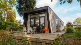 Lodge moderne appelé Just Nature entouré de verdure, terrasse avec chaises, journée ensoleillée.