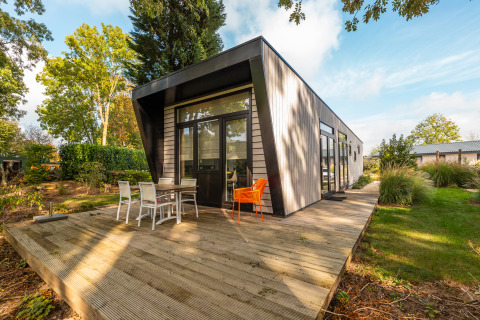 Moderne Lodge „Just Nature“ im Ferienpark Aan de Maas, Niederlande, mit Holzterrasse und Gartenmöbeln im Grünen.