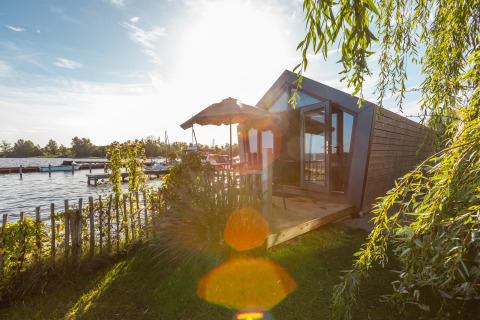Sunny lakeside view of Tiny House Havenzicht with a wooden deck, garden plants, and a clear sky.