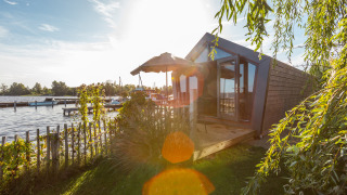 Sunny lakeside view of Tiny House Havenzicht with a wooden deck, garden plants, and a clear sky.