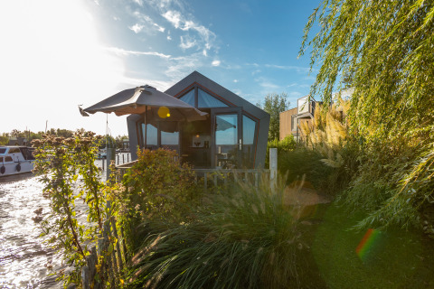 Moderna tiny house junto al agua con jardín y sombrilla en De Wiedense Meren, Países Bajos, al atardecer.