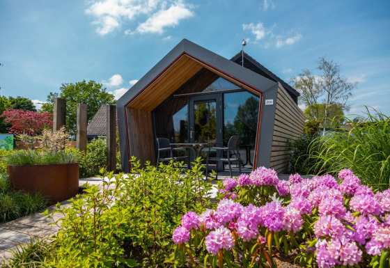 Tiny house moderna con grandi finestre, circondata da giardino verde e fiori rosa in primo piano.