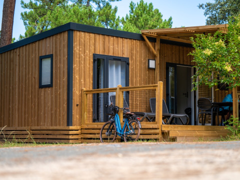 Alojamiento de glamping moderno de madera, con porche, bicicleta y mobiliario de exterior entre árboles.