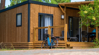 Alojamiento de glamping moderno de madera, con porche, bicicleta y mobiliario de exterior entre árboles.
