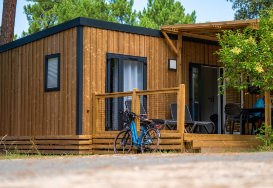 Alojamiento de glamping moderno de madera, con porche, bicicleta y mobiliario de exterior entre árboles.