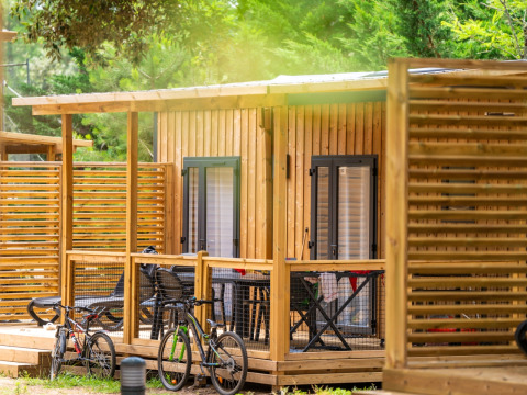 Alojamiento glamping con cabaña de madera, porche, dos bicicletas y naturaleza verde de fondo.