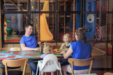 A family sits and eats at a table inside an indoor play park, with play structures behind glass windows.