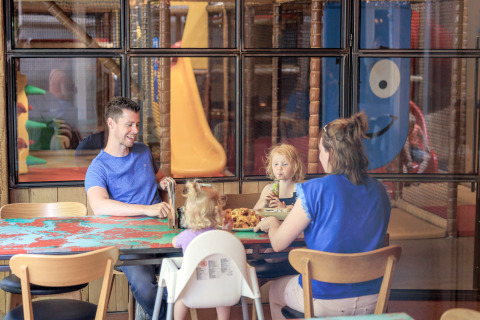 Een gezin zit te eten aan een tafel in een indoor speelpark, met speelstructuren achter glas.