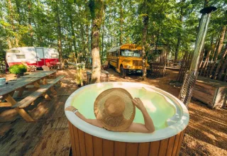Persona nella vasca idromassaggio con cappello al Chianti Glamping Resort, tra bosco, camper e scuolabus.