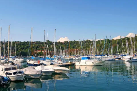 Barche e yacht ormeggiati nel porto turistico di Sistiana, Italia, circondati da colline verdi e cielo sereno.