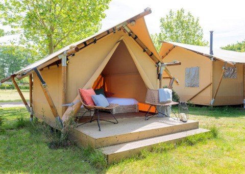 Tenda safari luminosa con terrazza in legno e sedie da esterno, situata su un prato con alberi verdi.