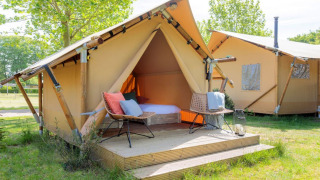 Tenda safari luminosa con terrazza in legno e sedie da esterno, situata su un prato con alberi verdi.