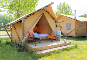 Ein Safari-Zelt mit Holzterrasse und Outdoor-Stühlen steht auf einer Wiese, umgeben von grünen Bäumen.