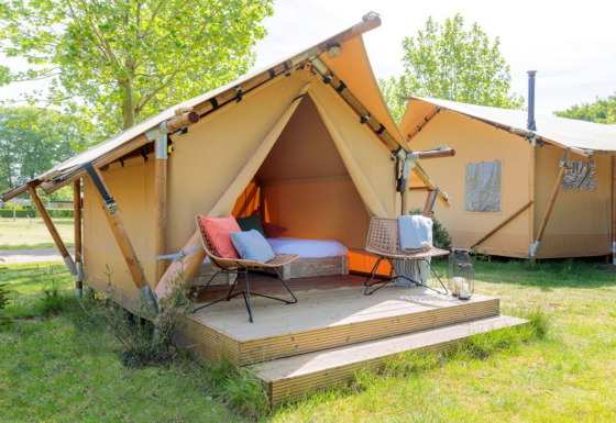 Lichte safaritent met houten terras en buitenstoelen, omgeven door gras en groene bomen in de natuur.