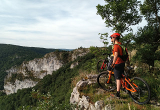 Mountain biker with orange bike on a cliff at Campsite Les 3 Cantons in Occitanie, France, overlooking lush valleys.
