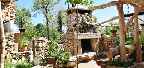 Stone fireplace and garden at Campsite Les 3 Cantons in Occitanie, France, surrounded by plants and wood.
