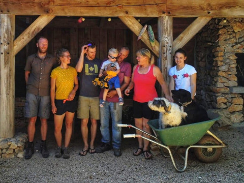 Foto di gruppo con sei adulti, un bambino e due cani in una carriola al campeggio Les 3 Cantons, Occitania, Francia.