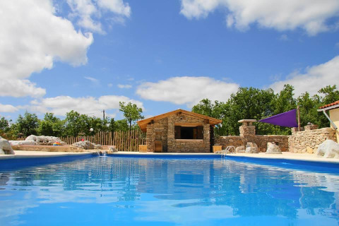 Piscina con edificio in pietra e alberi sullo sfondo al campeggio Les 3 Cantons in Occitania, Francia.