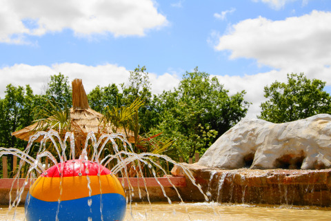 Fontana con palla colorata in un parco vacanze, tetto di paglia e alberi verdi sotto il cielo azzurro.