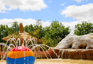 Colorful beach ball water fountain at a holiday park with thatched hut, rock feature, and lush green trees.