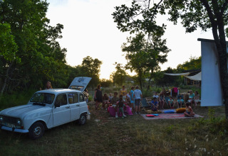 Outdoor cinema night with children and adults gathered near a vintage car at Les 3 Cantons campsite.