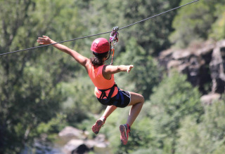 Persona su una zipline con casco rosso al campeggio «Les 3 Cantons» in Occitania, Francia, immersa nel verde.
